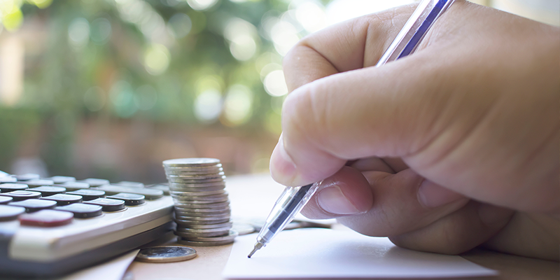 close up of hand writing, with calculator and pile of coins on desk