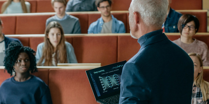 Lecturer holding laptop showing data graphic, talking to students in lecture theatre