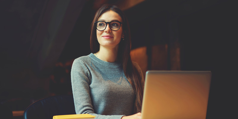 Female student sat at a table with laptop looking into the distance