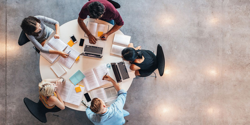 Five university students sat at a desk studying, with books open and laptops