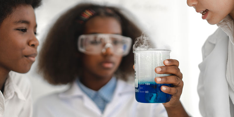 A school pupil is holding a science beaker with a smoking blue liquid inside. The pupil is wearing googles. Two other children are observing.