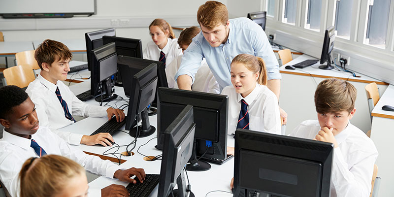 Secondary school pupils in an IT room. One child is getting help from a teacher.