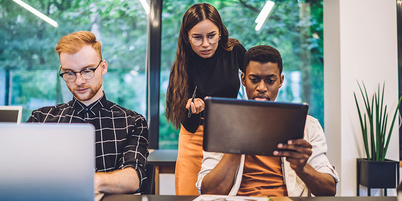 Three students - one is on a laptop, two are looking at a tablet