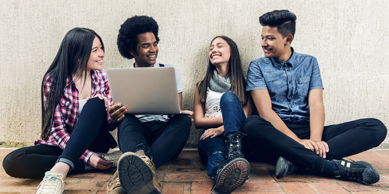 Four students sitting on the ground laughing, one with a laptop