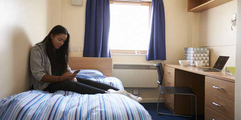 Student alone in their university residence, sat on the bed with a mobile phone