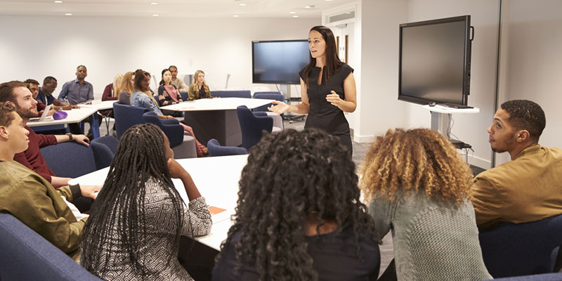 University lecture room with students gathered around tables, listening to a lecturer