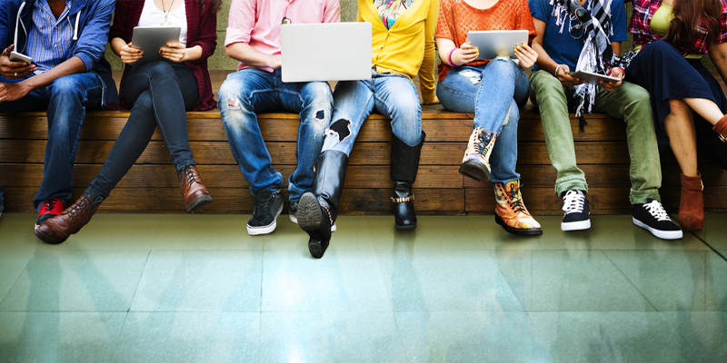 Row of students sitting on a bench, some with laptops